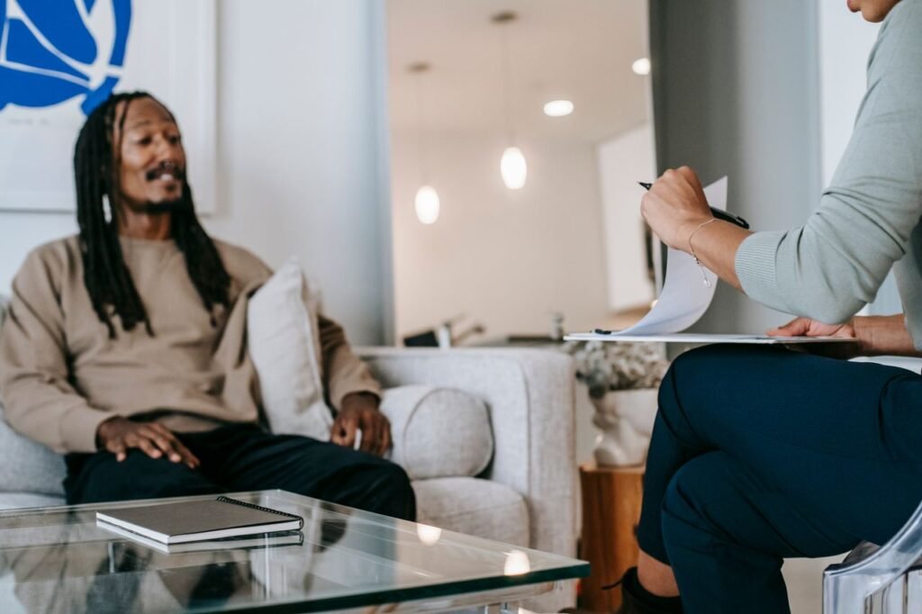pexels-photo-5699448-5699448 African American male with dreadlocks sitting on sofa in front of unrecognizable female therapist with clipboard during psychotherapy appointment in office